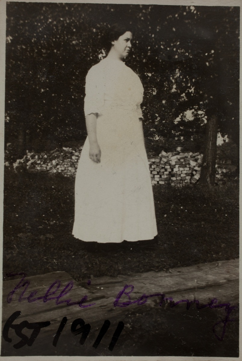 1911 ~ "Nellie Bonney Est 1911" standing in front of a brick pile in front of wooden walkway